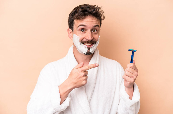 Young caucasian man wearing shaving foam and holding razor blade isolated on beige background smiling and pointing aside, showing something at blank space. - Photo, Image