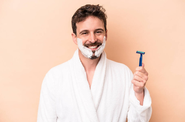 Молодий білий чоловік у піні для гоління і тримає лезо бритви ізольовано на бежевому фоні щасливий, усміхнений і веселий
. - Фото, зображення