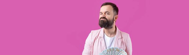 Retrato de un joven empresario satisfecho con una barba vestida con una chaqueta rosa que nos muestra billetes de dólar sobre un fondo rojo del estudio. Sabor, olor a dinero. - Foto, Imagen
