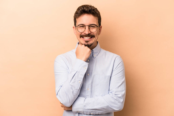 Young caucasian man isolated on beige background smiling happy and confident, touching chin with hand. - Foto, afbeelding