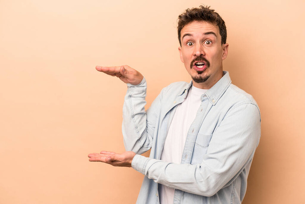 Young caucasian man isolated on beige background shocked and amazed holding a copy space between hands. - Foto, immagini