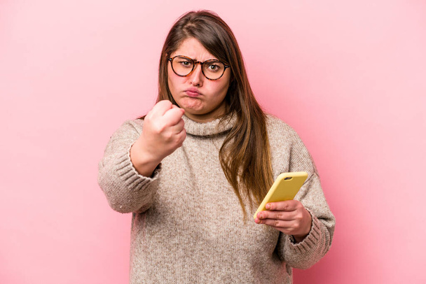 Jonge blanke overgewicht vrouw met een mobiele telefoon geïsoleerd op roze achtergrond tonen vuist naar camera, agressieve gezichtsuitdrukking. - Foto, afbeelding