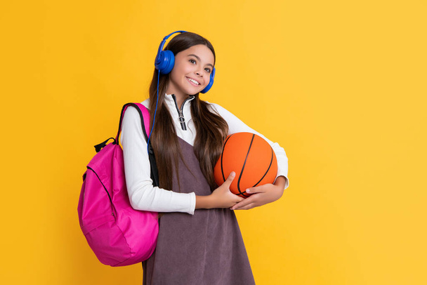 child smile in headphones with school backpack and basketball ball on yellow background. - Foto, imagen