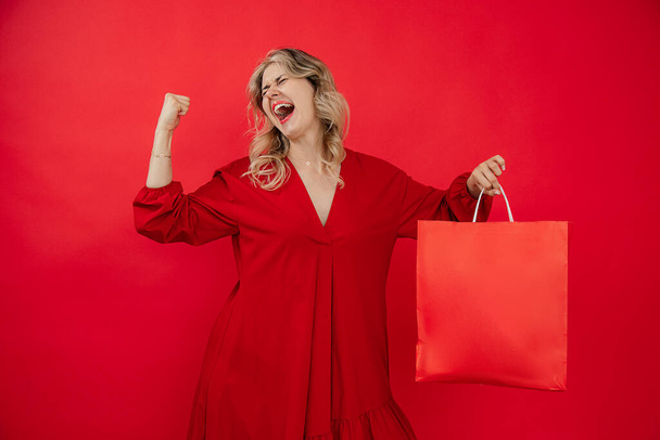  ενθουσιασμένοι ουρλιάζοντας WOW, πορτρέτο του ευτυχισμένη νεαρή χαριτωμένο γυναίκα ποζάρουν με τσάντες ψώνια πάνω από το κόκκινο φόντο. Ευτυχισμένη γυναίκα με τσάντες ψώνια απολαμβάνοντας στα ψώνια, κενό, αντίγραφο χώρου - Φωτογραφία, εικόνα