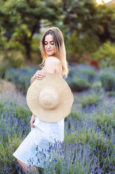 Jonge vrouw draagt een witte zonnejurk die haar hoed bedekt en naar beneden kijkt in een lavendelweide. Meisje genieten van lavendel geur zomer avond in het dorp. - Foto, afbeelding