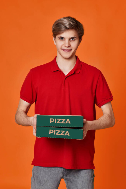 Pizza delivery man in red t-shirt holding cardboard boxes isolated on orange background - Zdjęcie, obraz