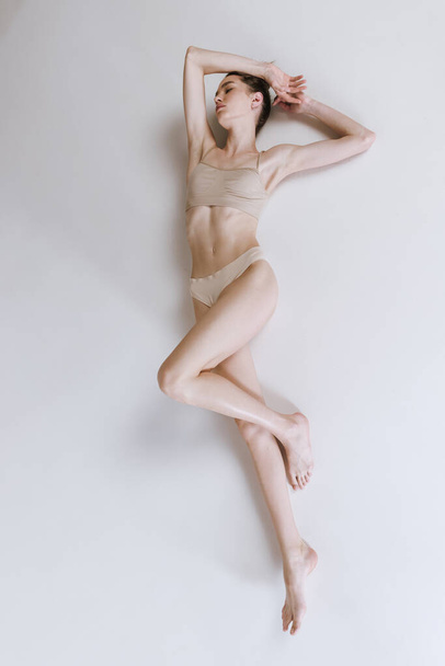 Portret van slanke gespierde vrouw liggend op de vloer in katoenen ondergoed, geïsoleerd poserend over grijze studio achtergrond. Concept van schoonheid, lichaams- en huidverzorging, gezondheid, spa, sport, dieet, cosmetica, reclame - Foto, afbeelding