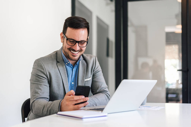 Fiatal mosolygós üzletember hálózatépítés online okostelefonon keresztül. Boldog férfi szakember a formálissal a laptopon dolgozik. Az íróasztalnál ül az irodában.. - Fotó, kép