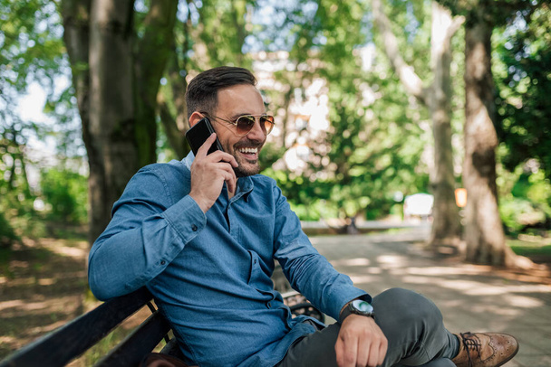 Onnellinen miesyrittäjä puhuu älypuhelimella. Hymyilevä nuori ammattilainen käyttää kännykkää istuessaan penkillä. Hänellä on viralliset vaatteet puistossa.. - Valokuva, kuva