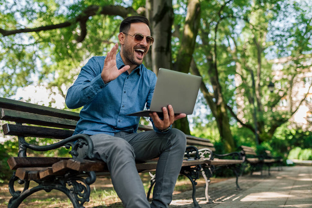 Gelukkige jonge zakenman zwaaiend op video oproep. Young professional is gebaren maken tijdens het netwerken via laptop. Hij zit op een bankje in een openbaar park in de stad.. - Foto, afbeelding