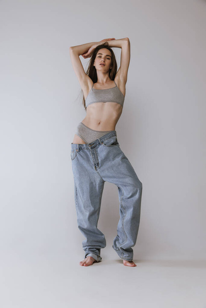 Portrait of young beautiful girl posing in top and oversized jeans isolated over grey studio background. Weight-loss. Concept of beauty, body and skin care, health, spa, sport, diet, cosmetics, ad - Fotó, kép