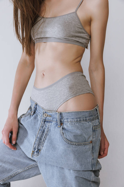Cropped image of slim female body in oversized jeans and underwear isolated over grey studio background. Weight loss. Concept of beauty, body and skin care, health, spa, cosmetics, ad - 写真・画像