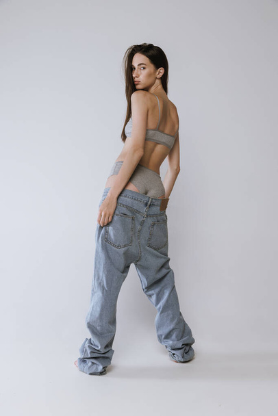 Schöne junge Frau posiert in großen Jeans und Baumwolltop isoliert über grauem Studiohintergrund. Model posiert in bequemer Unterwäsche. Konzept der Schönheit, Körper- und Hautpflege, Gesundheit, Wellness, Kosmetik, Werbung - Foto, Bild