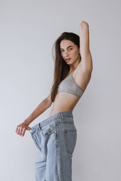 Portret van een aantrekkelijk jong meisje dat poseert in een gigantische oversized jeans geïsoleerd over een grijze studio achtergrond. Gewichtsverlies. Concept van schoonheid, lichaams- en huidverzorging, gezondheid, spa, cosmetica, reclame - Foto, afbeelding