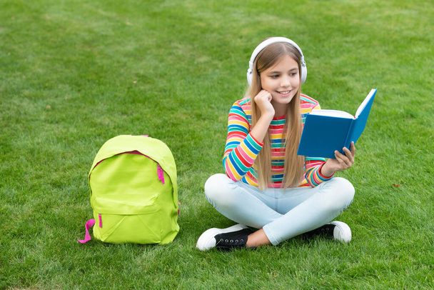 Happy teen girl reading book listening to headphones sitting on grass after school, homework. - Фото, изображение