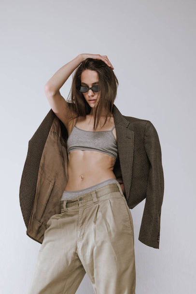 Portrait of stylish slim girl posing in sunglasses, pants and jacket isolated over grey studio background. Concept of beauty, fashion, style, lifestyle, body and skin care, health, cosmetics, ad - Photo, Image