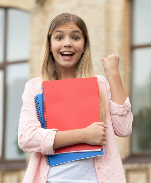 Happy pupil girl with books making winning gesture outdoors, back-to-school. - Foto, Imagem