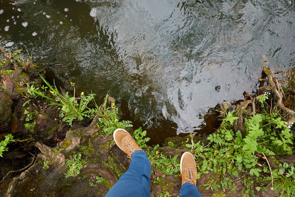 Homem em pé em terra em um parque florestal. Jeans azuis, botas castanhas. Rio. Plantas verdes, troncos de árvores, musgo, samambaia. Primavera, início do verão. Natureza, turismo, caminhadas, caminhada nórdica, estilo de vida saudável - Foto, Imagem
