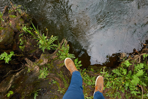 Mann steht an Land in einem Waldpark. Blaue Jeans, braune Stiefel. Fluss. Grüne Pflanzen, Baumstämme, Moos, Farn. Frühling, Frühsommer. Natur, Tourismus, Wandern, Nordic Walking, gesunder Lebensstil - Foto, Bild