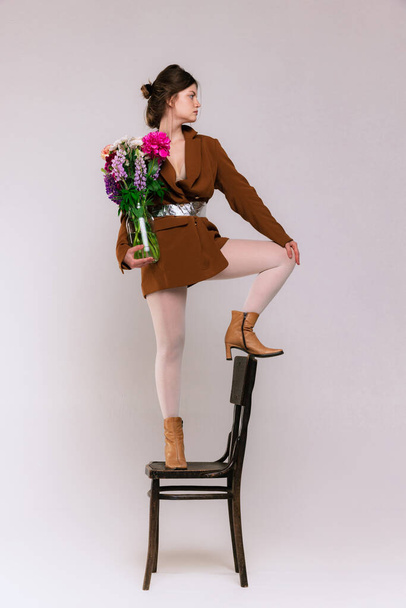 Portrait of stylish girl in vintage brown jacket, white tights, boots posing on chair with flowers isolated over grey background. Concept of retro fashion, art photography, style, queer, beauty - Photo, image