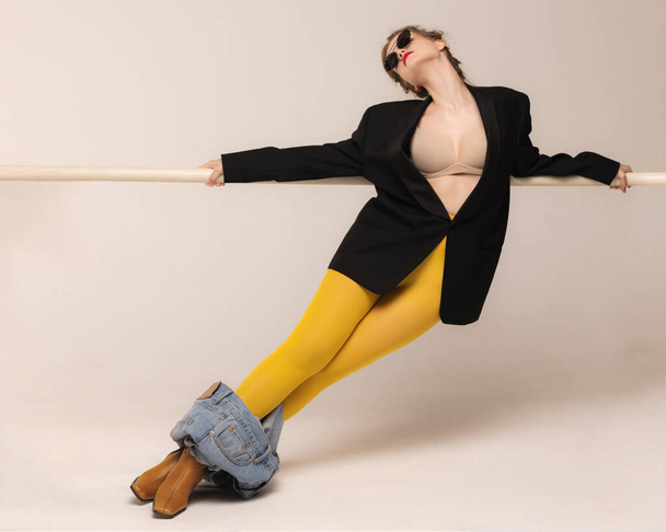 Portrait of young girl posing in yellow tights, dropped jeans and boots isolated over grey studio background. Feeling freedom. Concept of retro fashion, art photography, style, queer, beauty - Fotografie, Obrázek