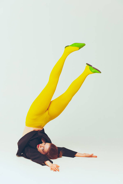 Film effect. Portrait of young stylish girl in yellow tight and black jacket posing with legs up isolated over grey studio background. Concept of retro fashion, art photography, style, queer, beauty - Photo, Image
