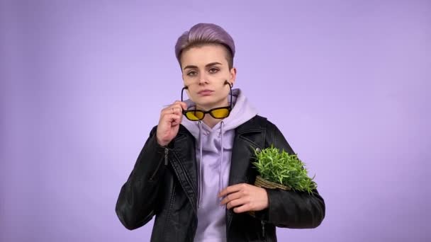 Fajna lesbijka z modną fryzurą w skórzanej kurtce, trzymając zieloną roślinę doniczkową, dając suchy cyniczny wygląd do kamery, zakładając okulary przeciwsłoneczne odizolowane na fioletowym tle w pomieszczeniach. Widok z przodu - Materiał filmowy, wideo