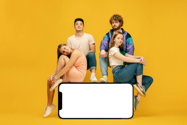 黄色の背景に隔離された空の白い画面を持つスマートフォンの巨大な3Dモデルに立って座っている若者のグループ,若者,新しいアプリやウェブサイト,広告のためのコピースペース,モックアップやデザイン - 写真・画像