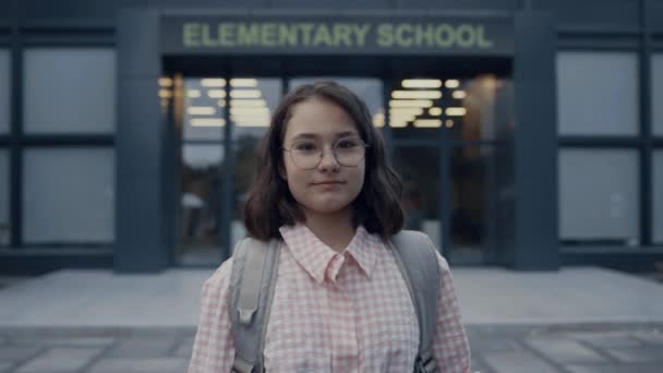 Serious school girl standing on schoolyard alone closeup. Cute shy teenager wearing glasses backpack posing in front building entrance after classes. Elementary age brunette looking camera outdoors. - Footage, Video