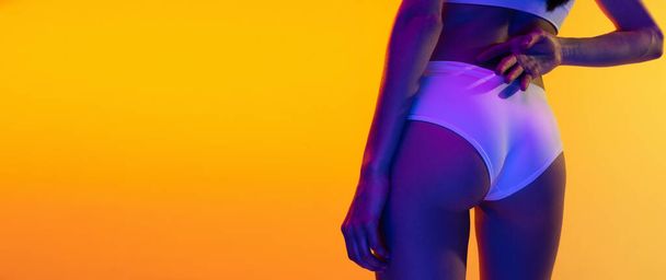 Cropped image of slim female body, buttocks isolated over yellow studio background in neon light. Anti-cellulite treatment. Concept of beauty, body and skin care, sport, health, spa, cosmetics, ad - Φωτογραφία, εικόνα