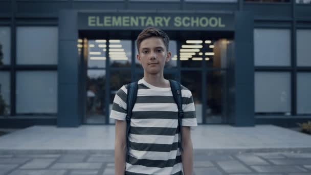 Close up of serious teen boy standing at empty schoolyard. Tensed schoolboy posing alone in front elementary school building. Portrait of pensive guy teenager wearing striped t-shirt with backpack. - Footage, Video