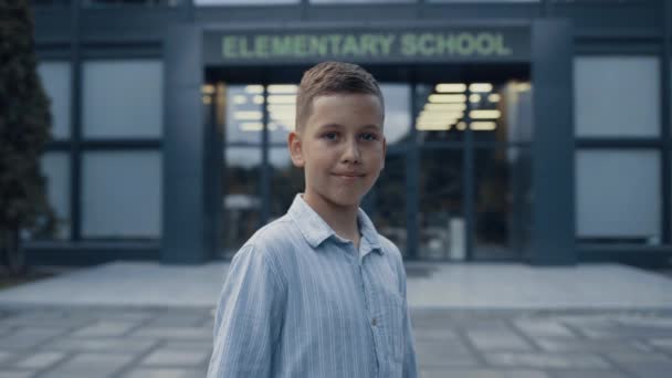 Aranyos, mosolygós, általános iskolás fiú, aki egyedül áll az iskola bejáratánál. Pihent tinédzser pózol a kampusz üvegajtajában, kamerát néz. Portré vidám iskolás fiú érzés boldog az első nap. - Felvétel, videó