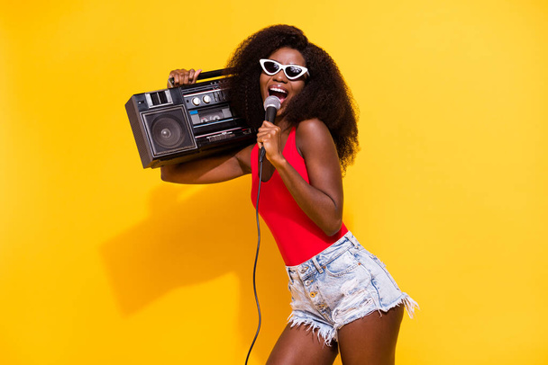 Photo of optimistic nice brunette lady hold boom box sing wear eyewear top shorts isolated on yellow color background. - Fotoğraf, Görsel