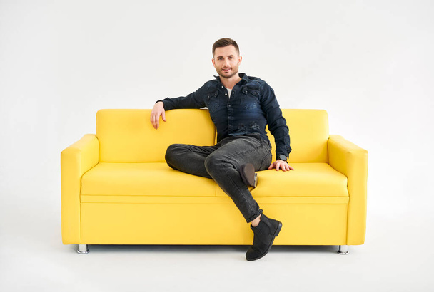 Young trendy man posing sitting on yellow sofa looking to camera. Relax concept - Φωτογραφία, εικόνα