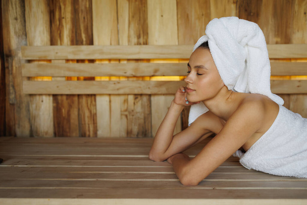 Young woman in white towel lying in wooden sauna - 写真・画像