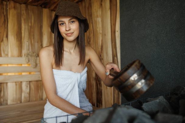 Retrato de uma menina bonita com uma concha de madeira na sauna derramando água em pedras quentes - Foto, Imagem