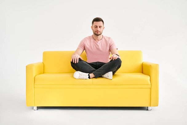 Handsome young man sitting in yoga lotus pose and meditating on comfortable yellow sofa over white background - Fotografie, Obrázek