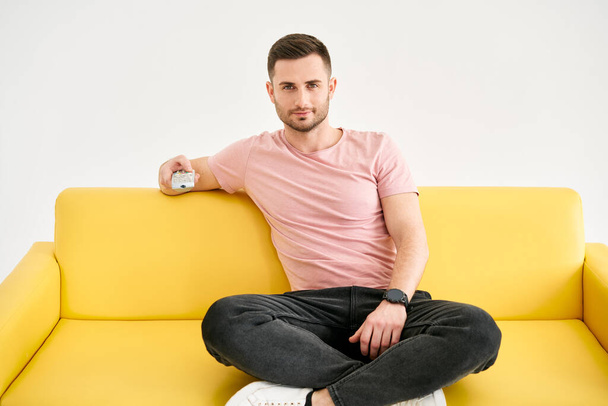 Young man watching TV with remote control sitting on comfort sofa over white background. Male looking to camera holding TV remote control - Fotó, kép