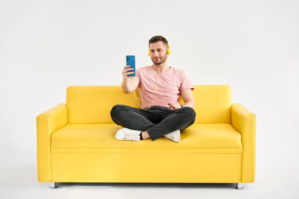 Relaxed young man wear headphones listen music by his mobile phone while sitting on yellow sofa over white background - Foto, Imagen