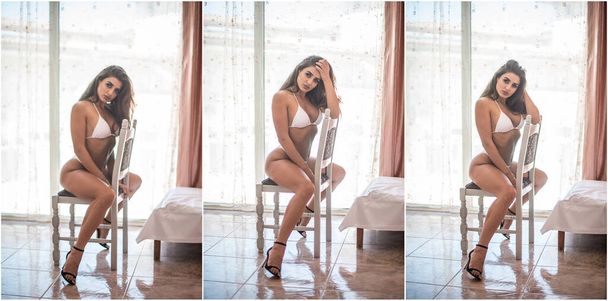Attractive sexy brunette in white lingerie  posing provocatively, on chair in front of a window . Portrait of sensual woman with long hair, white bikini and bra in classic boudoir scene in studio - Φωτογραφία, εικόνα