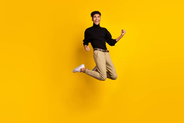 Photo of funny lucky dark skin guy dressed black pullover jumping rising fists isolated yellow color background. - Foto, Imagem