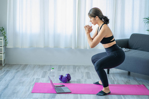 Mujer asiática sonriente haciendo estiramiento de hombros clase de yoga en línea desde la computadora portátil en casa en la sala de estar. Autoaislamiento y ejercicio en casa con un sofá y un ordenador portátil al lado. - Foto, Imagen
