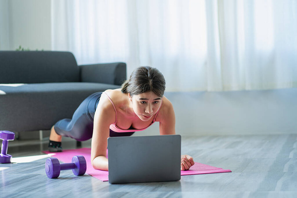 Glimlachende Aziatische vrouw doet schouder stretching yoga online klasse van laptop thuis in de woonkamer. Zelfisolatie en lichaamsbeweging thuis met een bank en laptop ernaast. - Foto, afbeelding