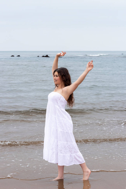 молода брюнетка, піднімаючи руки на пляжі в білій сукні
 - Фото, зображення