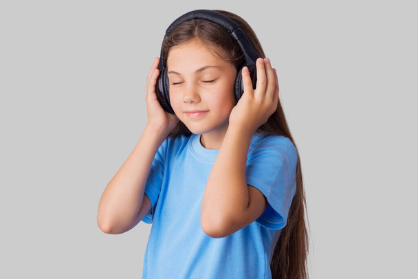 Kleines Mädchen im blauen T-Shirt hört Musik mit drahtlosen Kopfhörern und fühlt sich wohl. Studioaufnahme eines kleinen Schulmädchens vor grauem Hintergrund - Foto, Bild