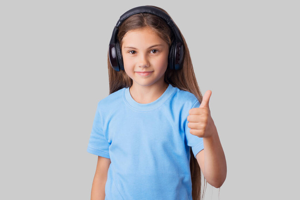 Petite fille souriante écoutant de la musique et montrant les pouces sur un fond gris. Écolière aime écouter de la musique à l'aide de grands écouteurs studio - Photo, image