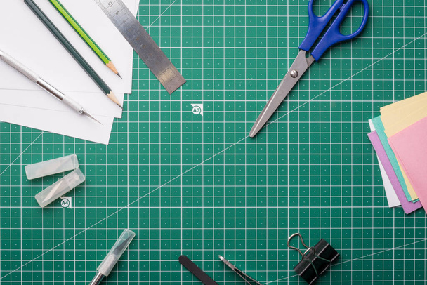 Scalpel, pincet, liniaal, paperclip, doos met messen, gekleurd papier, potloden, schaar op snijmat. Verschillende kantoorbenodigdheden op groene professionele snijmat  - Foto, afbeelding