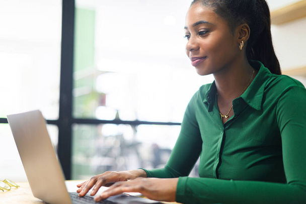 Amistosa y positiva empleada afroamericana o estudiante usando laptop, mujer de negocios multirracial mirando la pantalla, escribiendo, respondiendo a correos electrónicos sentados en una oficina contemporánea - Foto, Imagen