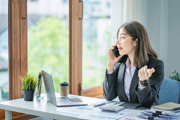Femme d'affaires asiatique travaillant dans le bureau avec des papiers et tenant un stylo à l'aide d'un ordinateur portable assis sur une chaise à la maison moderne studio mobile travailleur contact avec les clients - Photo, image