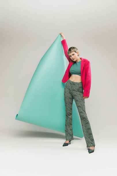 Portrait of young girl in bright jacket and vintage pants posing with paper piece isolated over grey studio background. Concept of retro fashion, art photography, style, queer, beauty - Fotó, kép